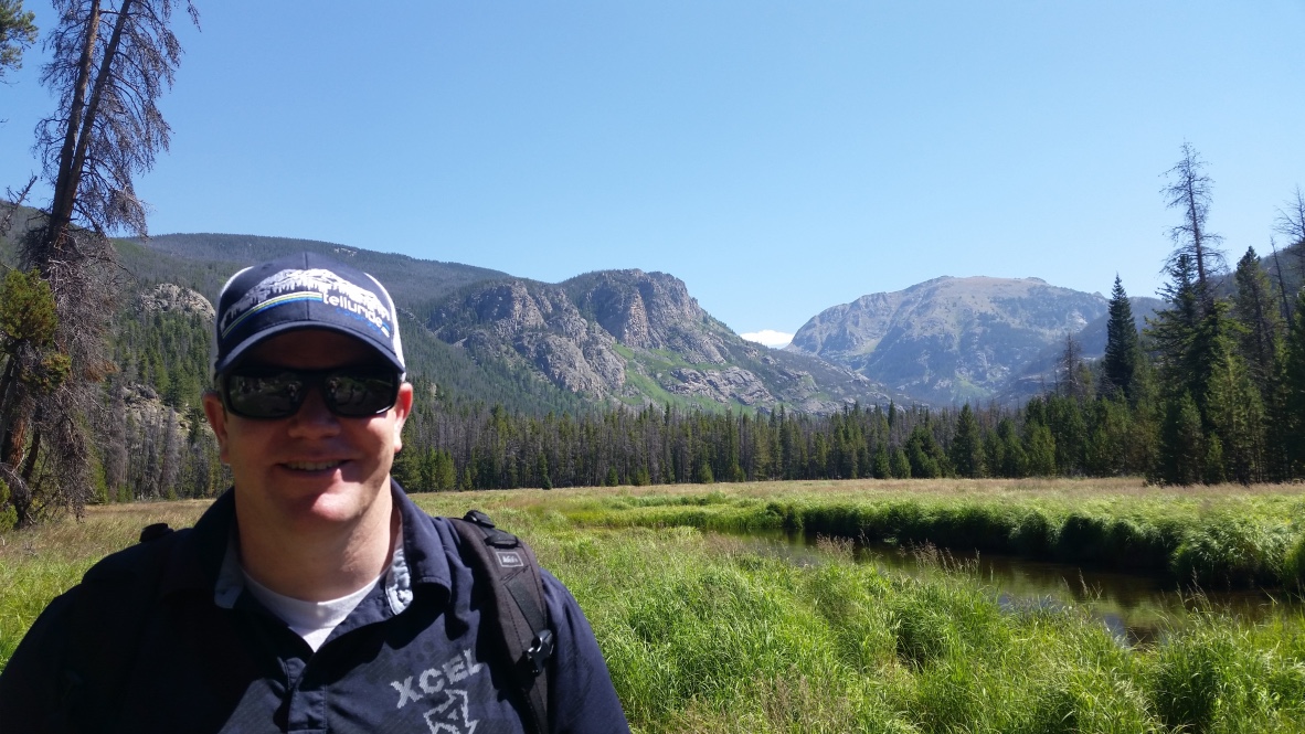 A man smiling in front of a river and mountains.